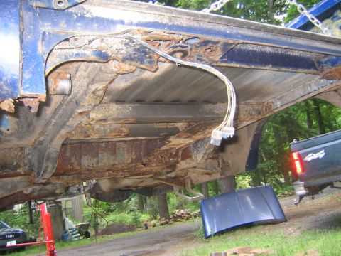 1978 Ford Bronco - Underside view of a blue pickup bed hoisted on a chain lift, showing heavy rust perforation through the bedside, floor ribs and cross membe…