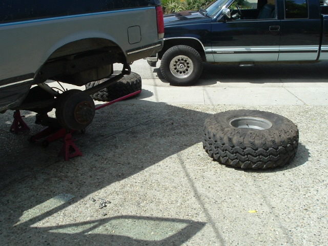 Blue and silver Bronco on jack stands with rear wheel off, spare tire on driveway