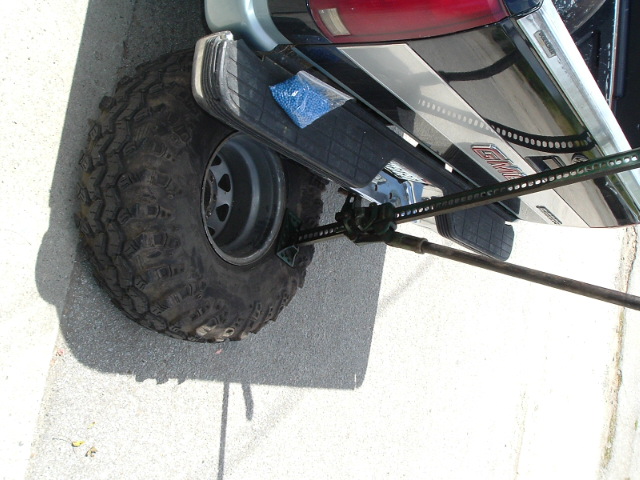 Mud-terrain tire on black steel wheel strapped beneath the chrome rear bumper of a GMC pickup