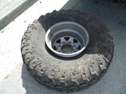 1989 Ford Bronco - A large off-road tire with a tread design is mounted on a silver rim, resting on an asphalt surface.