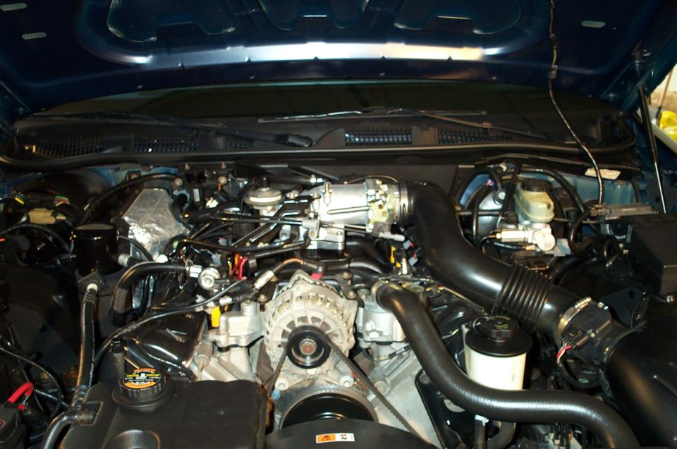 Engine bay of a vehicle showing alternator, air intake tube and accessory belt under a blue hood