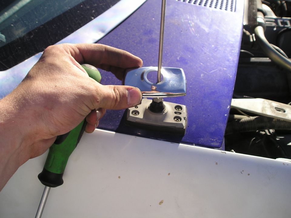 Hand using green-handle screwdriver to pry a clip near a blue painted body panel and windshield