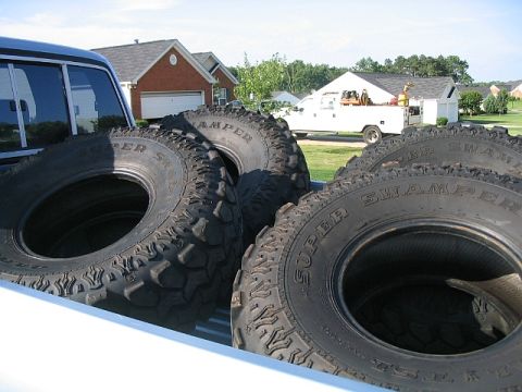 1986 Ford F-150 - A close-up view of off-road tires loaded into a truck bed, with houses and another truck visible in the distance.