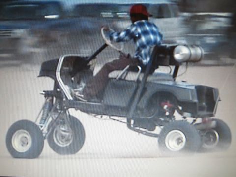 1990 Ford Bronco - rider in a plaid shirt and red cap pilots a low tube-frame sand buggy fitted with a cylindrical tank.