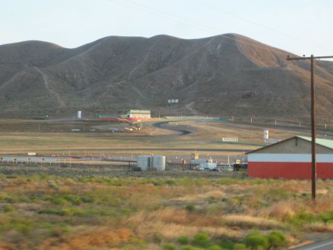 1990 Ford Bronco - distant view across scrubland of a racetrack, infield buildings and a red-and-white barn beneath dry mountains.