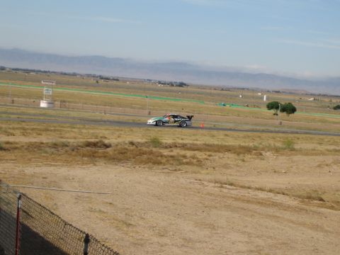 1990 Ford Bronco - winged race car rounds a road course set in a flat dry valley with distant mountains and trackside signage.