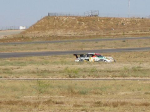 1990 Ford Bronco - white-and-dark liveried race car with a rear wing on a road course beside a dirt embankment with spectators above.