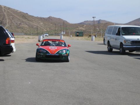 1990 Ford Bronco - red-and-green Mazda Miata race car numbered 55 sits in a paved paddock between an SUV and a white van.