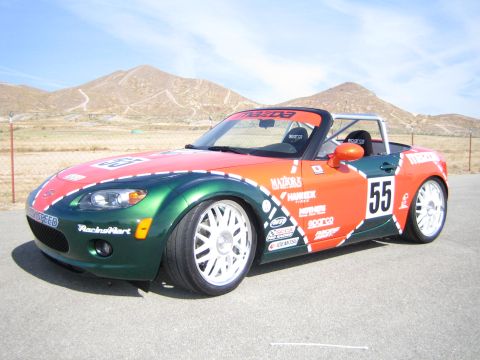 1990 Ford Bronco - green-and-orange liveried Mazda Miata roadster race car numbered 55 with white wheels parked before dry hills.