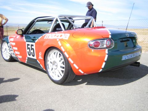1990 Ford Bronco - Rear three-quarter view of an orange-and-green Mazda Miata race car numbered 55 with a man standing behind it.