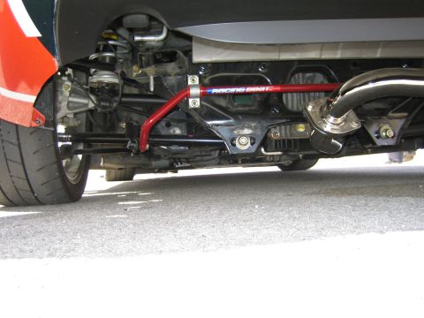 1990 Ford Bronco - Underbody view of a car showing a red Racing Beat sway bar, suspension links and exhaust routing on pavement.