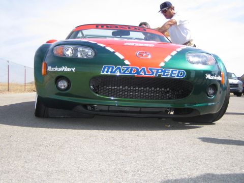 1990 Ford Bronco - green Mazda MX-5 roadster wearing Mazdaspeed and Racing Hart decals sits on a sunny paddock as men work near the cockpit.