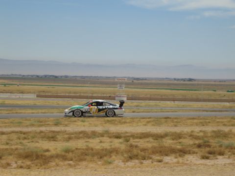 1990 Ford Bronco - liveried sports car with a rear wing speeds along a racetrack against a backdrop of dry grass and hazy mountains.
