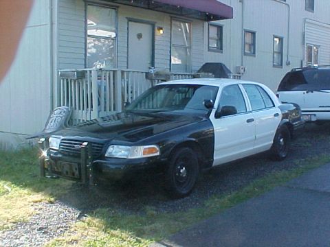 1999 Ford Crown Victoria - A two-tone police car is parked next to a building with another vehicle behind it.