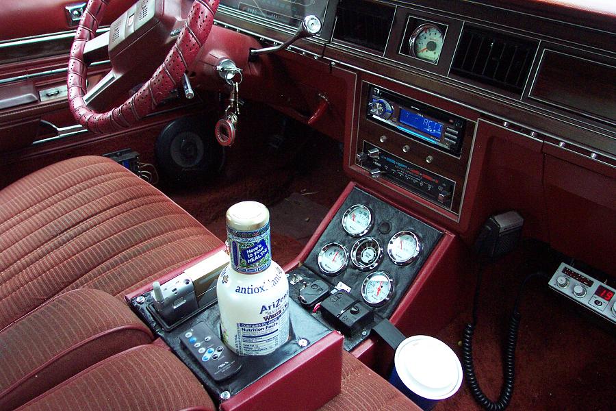 Car interior showing red seats, steering wheel, dashboard with radio, and aftermarket gauges in the center console. Bottle of Arizona antiox is on the console