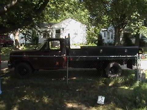 1986 Ford Bronco - A dark-colored flatbed truck is visible parked in a grassy area behind a chain-link fence, with houses and trees in the background.