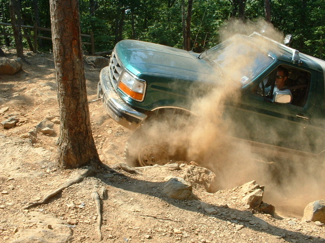 Green Ford truck kicking up dust on a rocky trail, driver visible