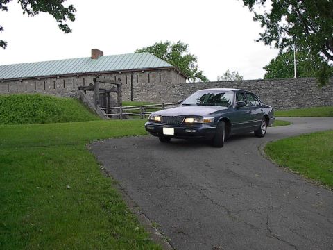 1985 Mercury Grand Marquis - A dark green 1996 Mercury Grand Marquis is seen driving on a road near a historic stone structure.