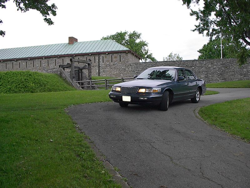 Dark green sedan, 1996 Mercury Grand Marquis, driving on a paved road in front of a stone building