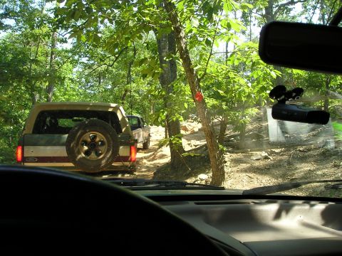 1996 Ford Bronco - Two SUVs navigate a wooded trail, kicking up dust on the path ahead.