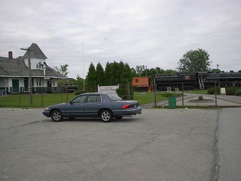1985 Mercury Grand Marquis - A blue sedan is parked in a lot next to a historic train station and railway museum.