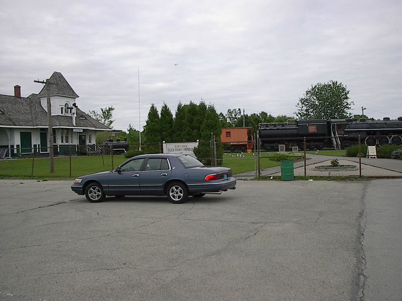 Blue-gray sedan parked side-on outside a railroad museum with steam locomotive