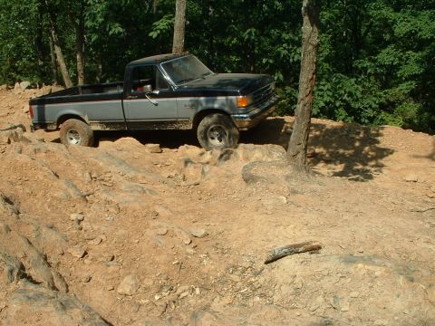 1985 Ford Bronco - A two-tone Ford F-Series truck navigates a challenging, rocky off-road path surrounded by trees.