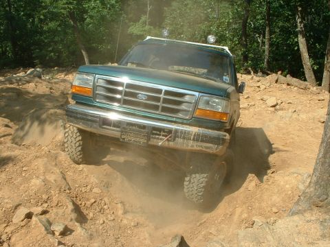 1985 Ford Bronco - Head-on view of a green Ford 4x4 with chrome grille and Ford oval dropping through a dusty rock-and-dirt trail, front tires flexed over the…