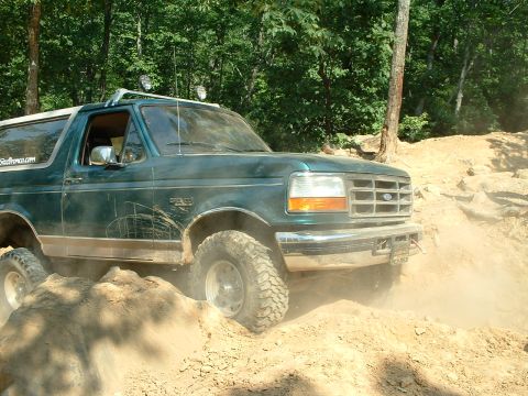 1985 Ford Bronco - A green Ford Bronco navigates a challenging off-road terrain with dust kicking up.