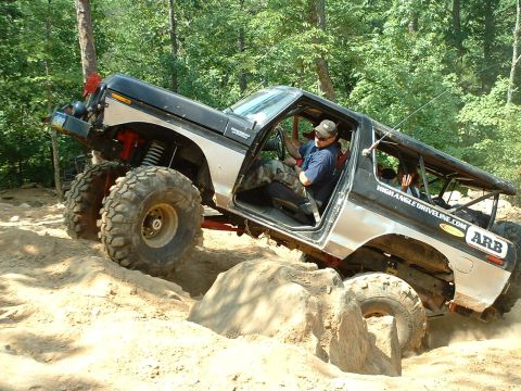 1985 Ford Bronco - A doorless, roof-less full-size Ford Bronco in silver and dark blue with a full roll cage climbs a rocky dirt hill, front axle articulating…