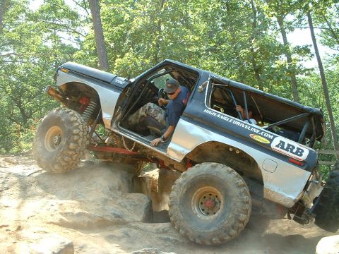 1985 Ford Bronco - A blue-and-white full-size Ford Bronco with no doors, a full internal roll cage and topless rear crawls over a rock ledge on a wooded trail…