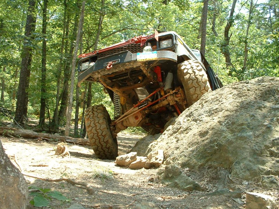 Modified off-road vehicle with large tires climbing a rock face in a wooded area