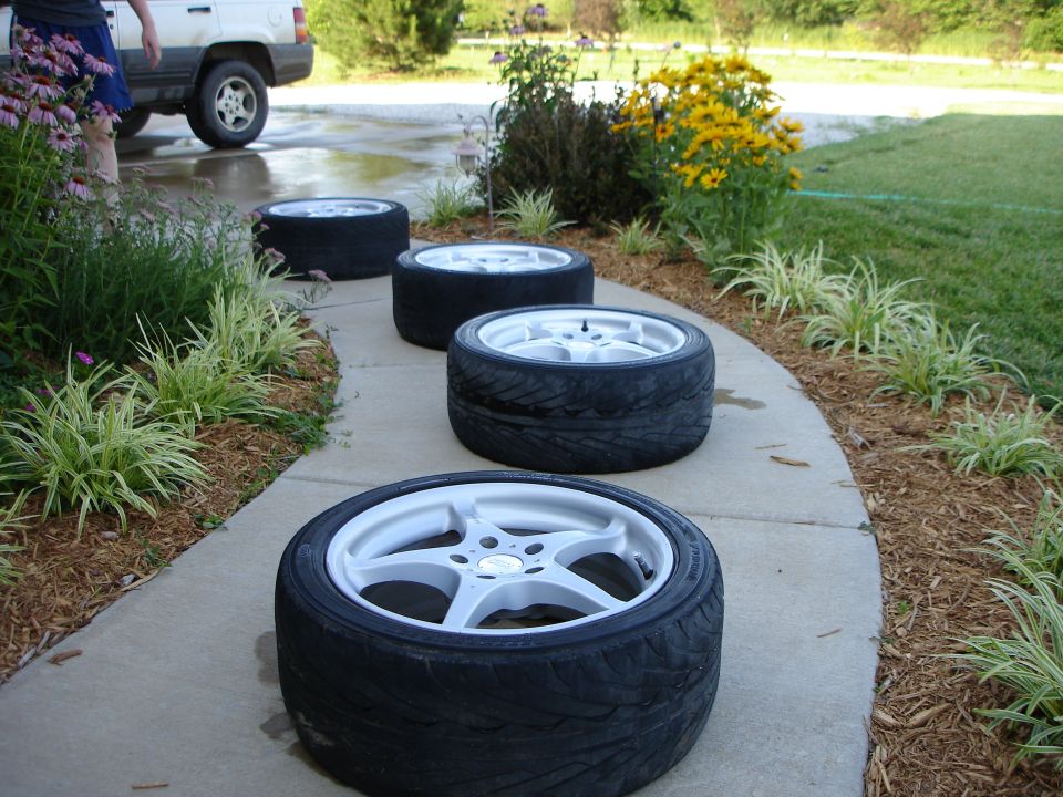 Four white five-spoke alloy wheels with tires lined up on garden walkway