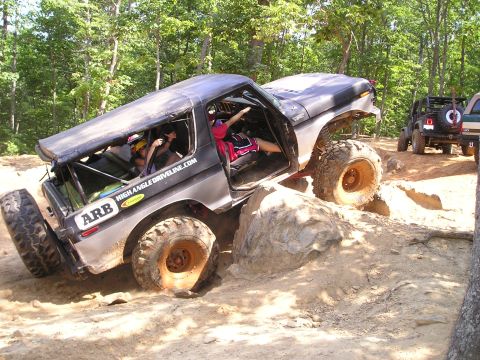 1996 Ford Bronco - A modified Ford Bronco attempts to clear large rocks on an off-road trail, with another Jeep visible in the background.