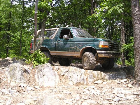 1996 Ford Bronco - A green 1996 Ford Bronco navigates a rocky off-road path surrounded by trees.