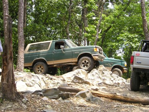 1996 Ford Bronco - Two Ford Broncos navigate a rocky off-road trail in a wooded area.