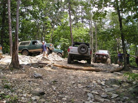 1996 Ford Bronco - Multiple off-road vehicles traverse a challenging, rocky path through a dense forest.