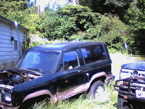 1987 Ford Bronco II - A 1987 Ford Bronco II sits in overgrown grass next to another vehicle, with a house and trees in the background.