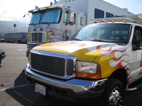 1990 Ford Bronco - white pickup with yellow-and-red flame graphics and a California plate sits in a lot beside a flame-painted cabover semi truck.