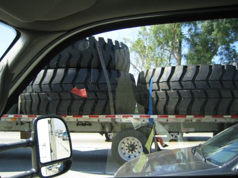 1990 Ford Bronco - Massive off-road tires strapped to a flatbed trailer are framed through the rear side window of a passing vehicle.
