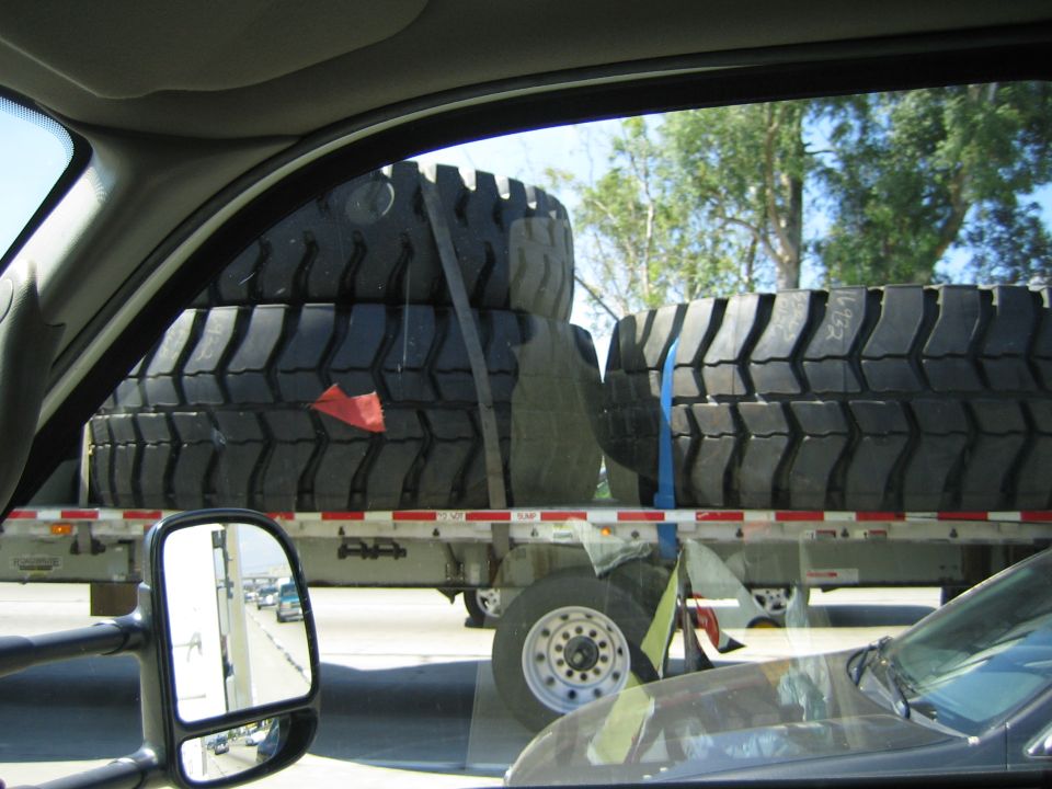 Oversized off-road tires on a flatbed trailer viewed through a vehicle's rear side window