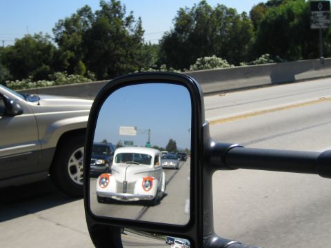 1990 Ford Bronco - tow mirror reflects a pale vintage hot rod coupe with painted fender accents cruising on the highway behind.