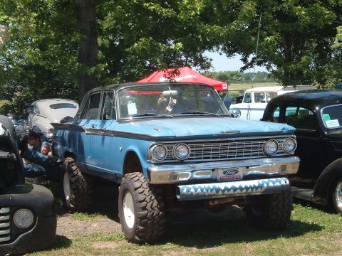 1994 Ford Bronco - A modified classic Ford sedan with oversized tires sits parked outdoors, with other vehicles and tents in the background.
