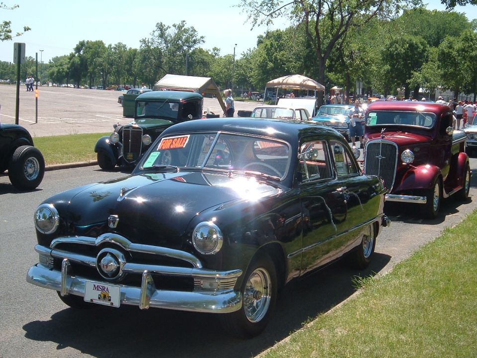 Black 1950 Ford sedan with a 'For Sale' sign in the window, parked on a street with other classic cars