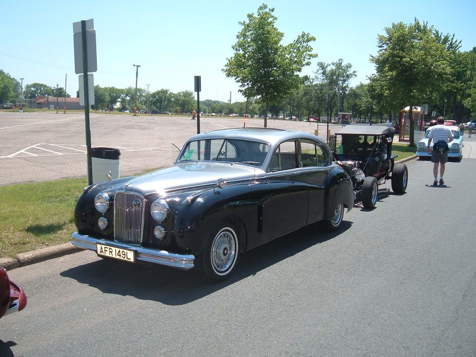 Black and silver sedan with a license plate reading AFR 149L, parked on asphalt next to a hot rod