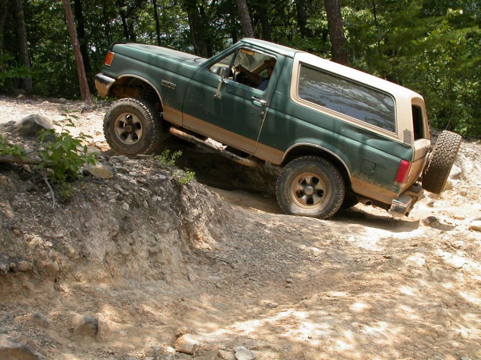 Green Ford Bronco with a tan top on a dirt trail, flat front tire, spare tire mounted on rear