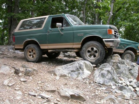 1990 Ford Bronco - A green Ford Bronco navigates a rocky off-road trail, with another Bronco visible behind it.
