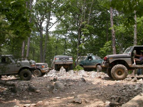 1990 Ford Bronco - Several off-road vehicles navigate a rocky, wooded area, with one vehicle prominently a driver.