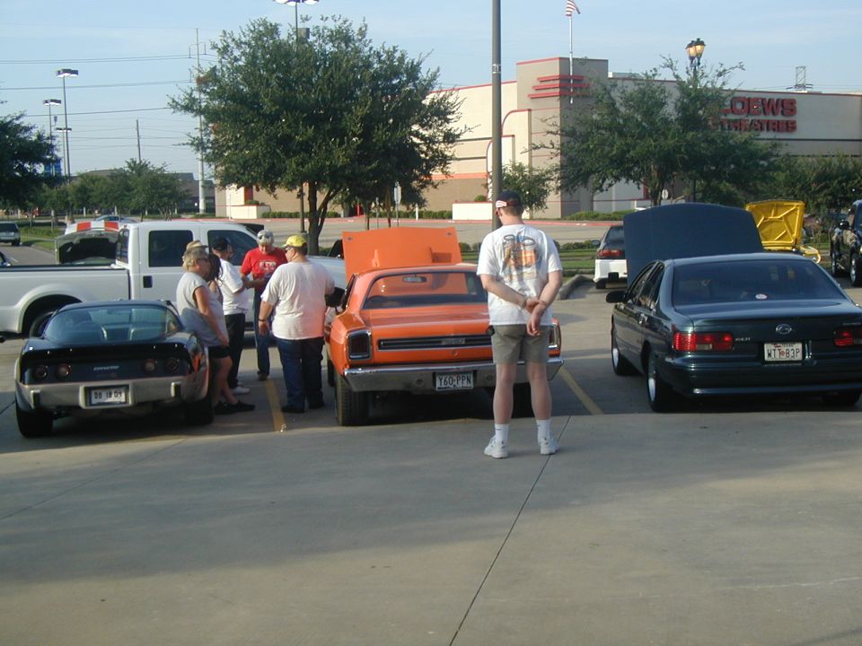 People gathered around cars at a parking lot meet, including a black Corvette, an orange classic car, and a dark sedan
