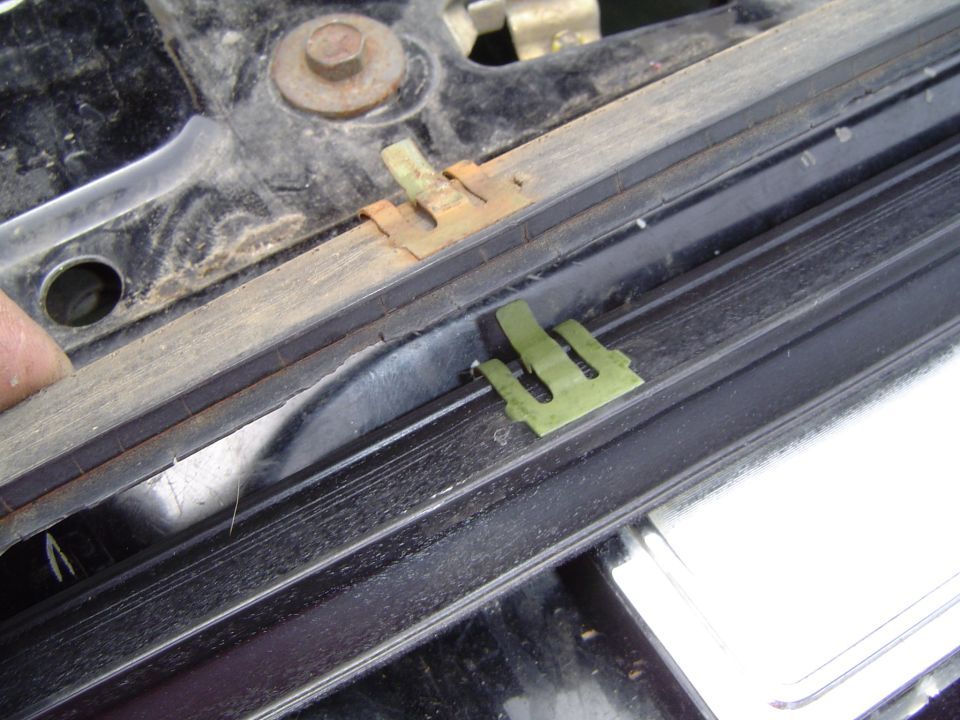 Green plastic clip and rusty bracket in a black window/door channel, finger at left.